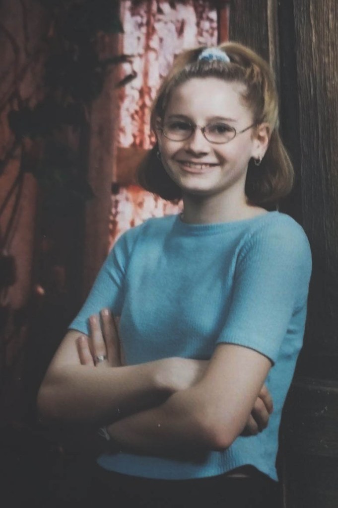 An 11-year-old Dezirae Ritchie stands for a school picture with her arms folded. She wears glasses, is smiling and wears a scrunchie in her hair.  
